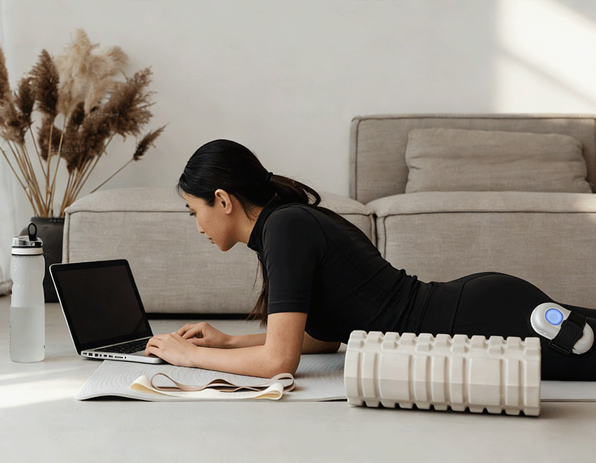 Woman lies on a mat using a laptop while wearing a SweetMyo controller on her thigh in a home care setting.