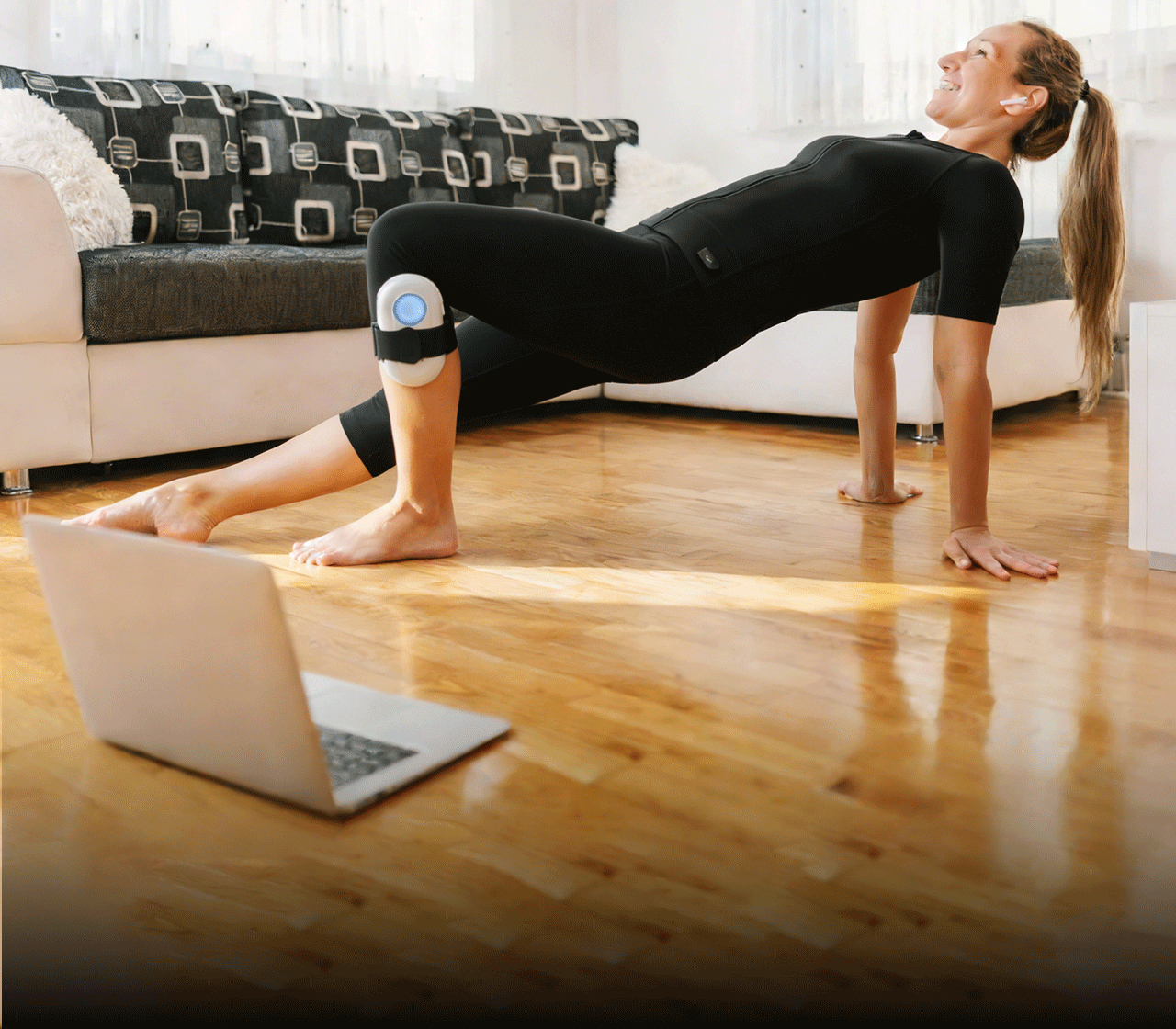 Woman in a black SweetMyo training suit performs a bridge exercise at home with a thigh-mounted controller and laptop nearby.