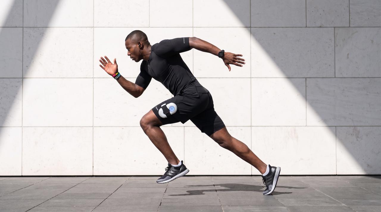 A fit man wearing a full-body EMS suit sprints powerfully outdoors against a white wall, showcasing how electric muscle stimulation enhances athletic performance and supports intense cardiovascular training sessions.