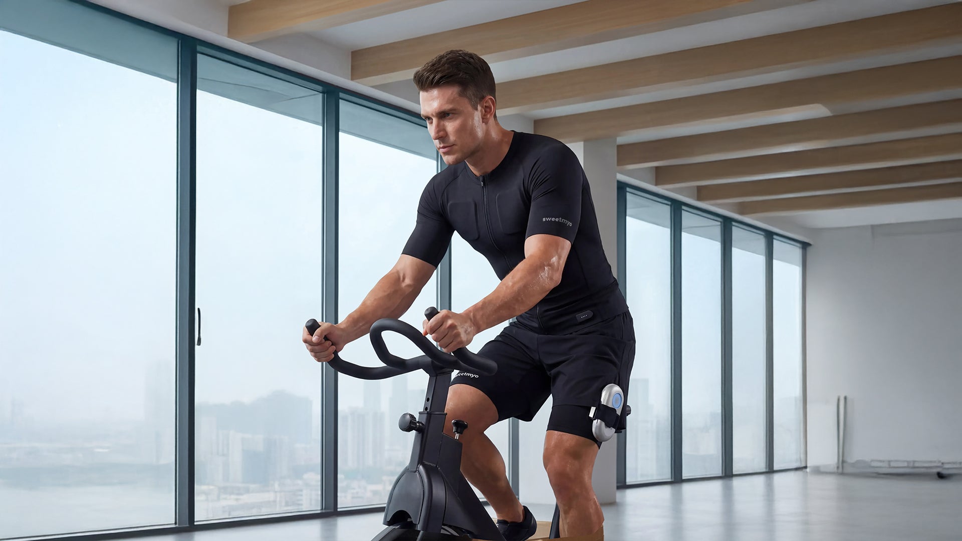 A fit man wearing a black full-body electrical muscle stimulation suit performs a cycling workout on a stationary bike inside a modern, well-lit apartment with large city-view windows.