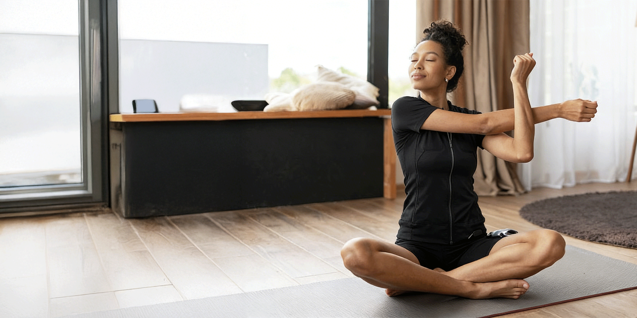 Woman wearing a black SweetMyo training suit stretches on a yoga mat in a bright home workout space.