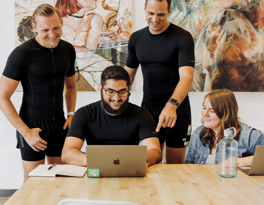 Team members gather around a laptop while some wear black SweetMyo training suits in a workplace wellness setting.