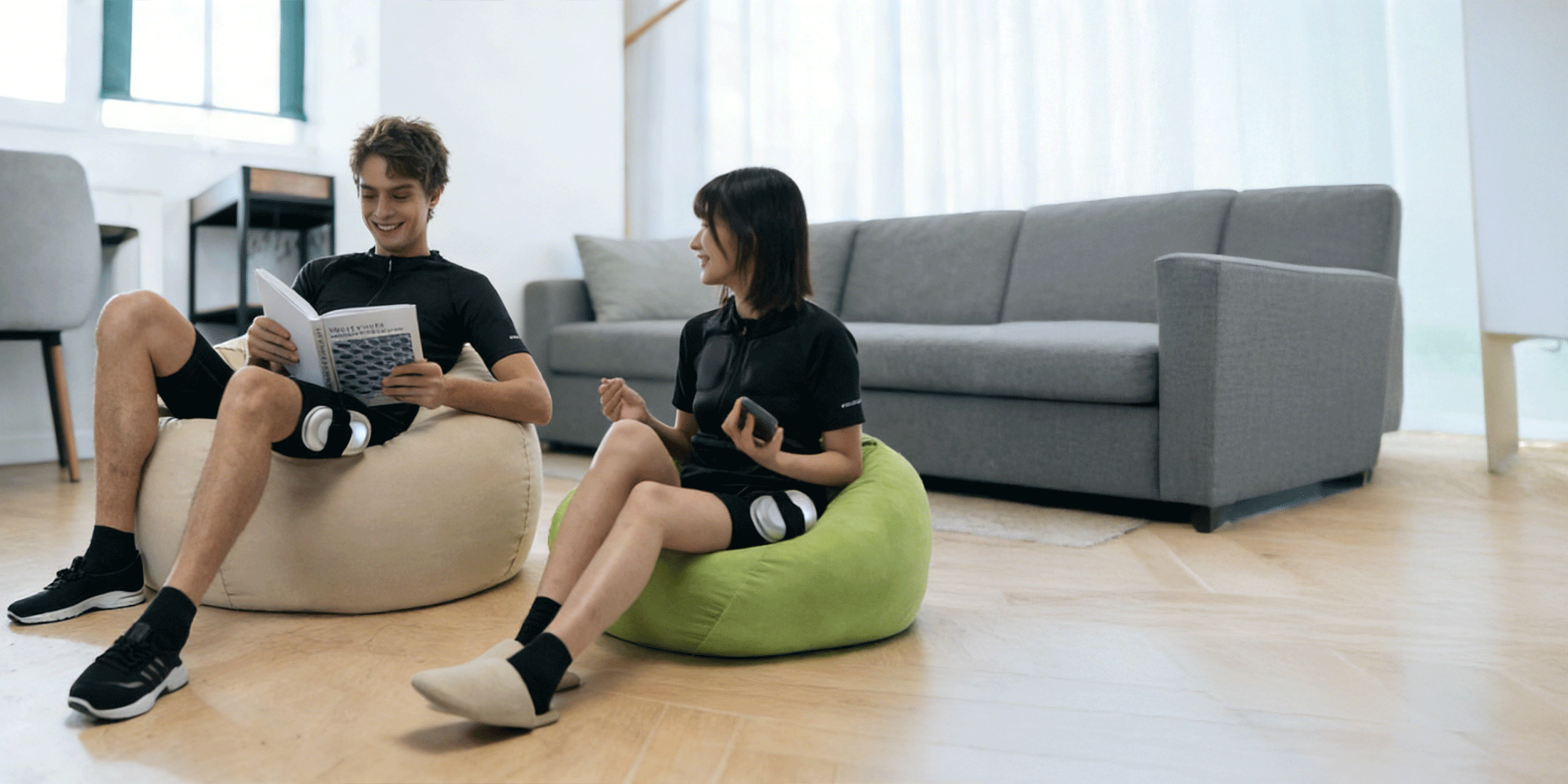 Two people wearing SweetMyo training suits relax on beanbags at home with wearable controllers attached to their thighs.